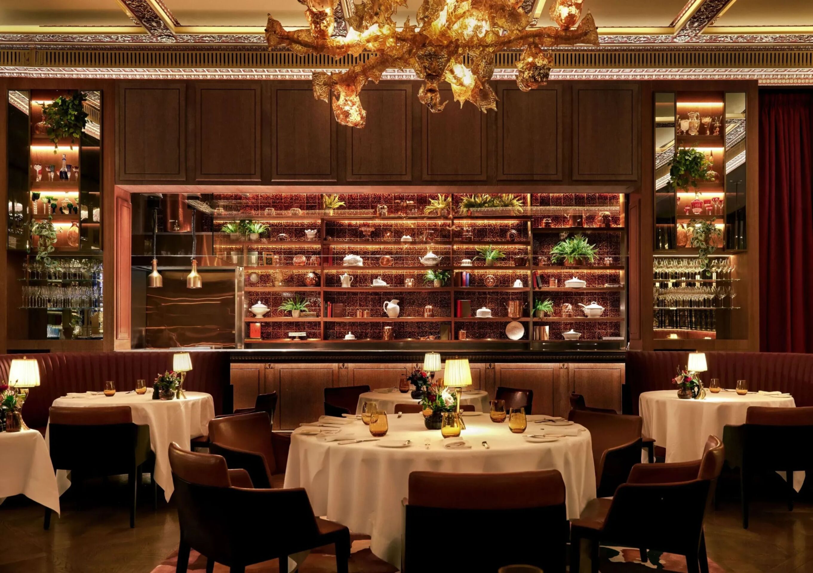 Interior of The Grill at The Dorchester, showcasing round dining tables set with white linens, warm ambient lighting, curved banquette seating and a dramatic golden chandelier suspended above the dining room.