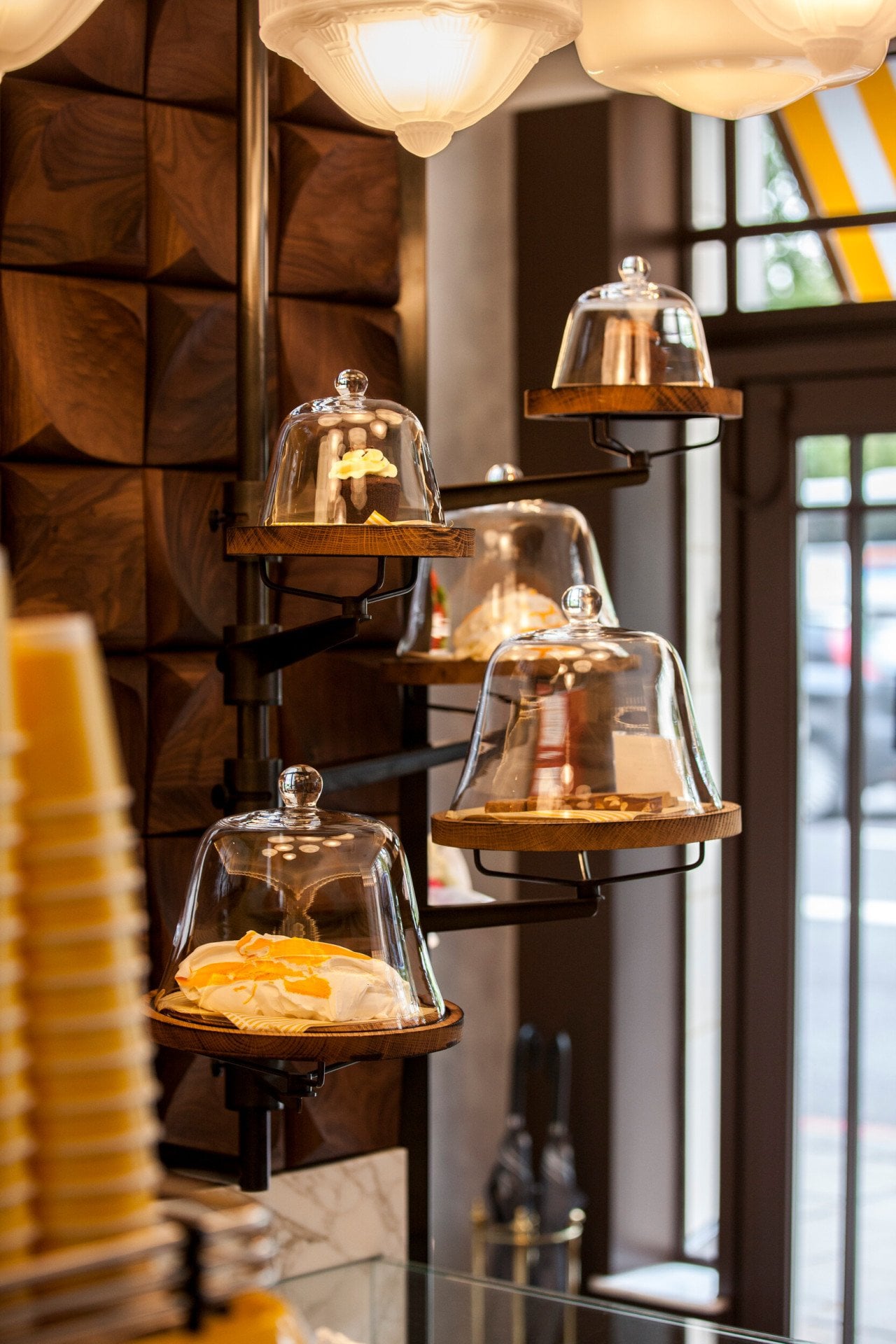 Display of freshly prepared pastries at Parcafé at The Dorchester, showcasing cakes and sweet treats arranged on tiered wooden shelves beneath glass cloches, illuminated by warm lighting and set against rich wood panelling.