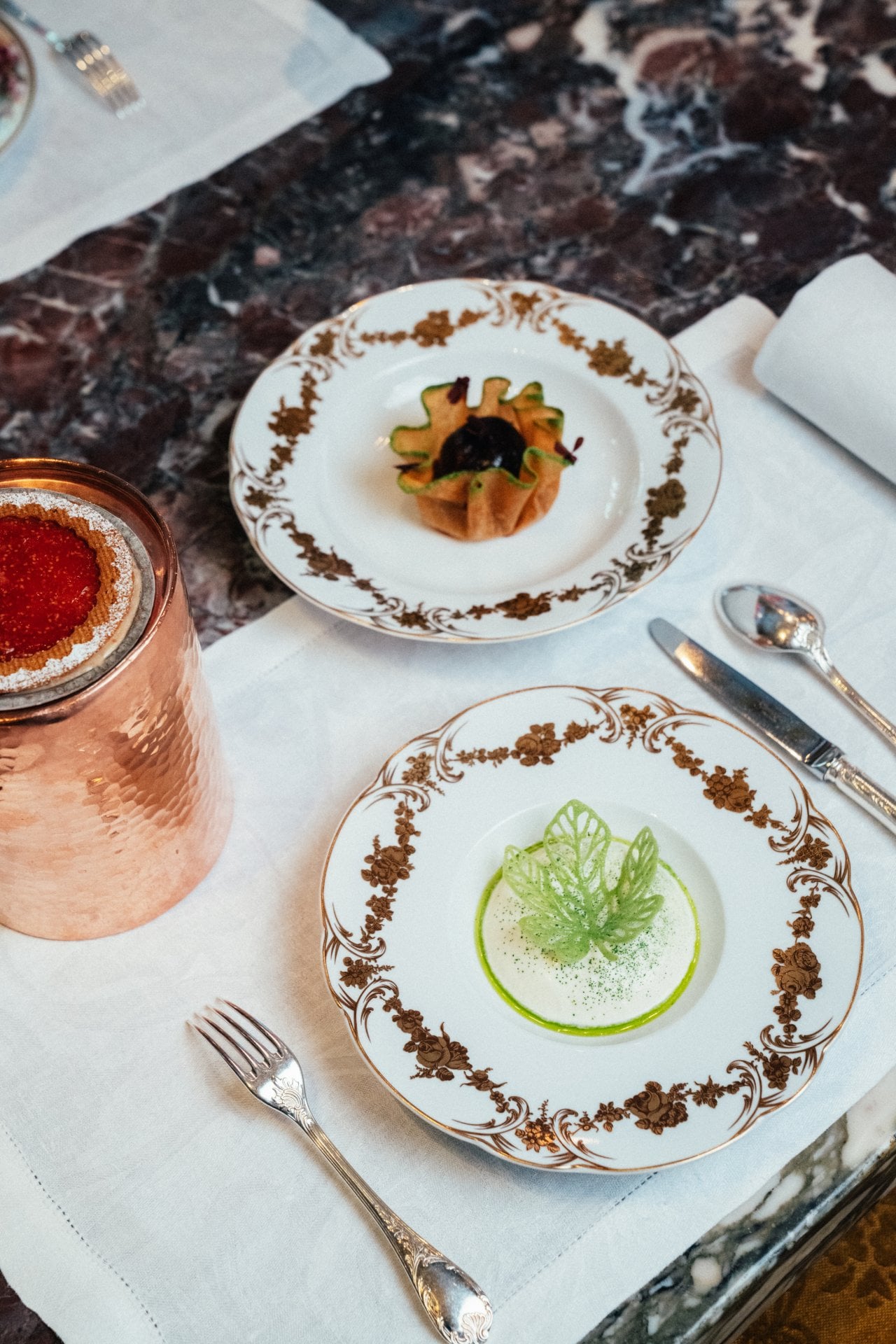 Elegant dining setup with decorative plates, small botanical-shaped entrees, silverware, and a copper tumbler.