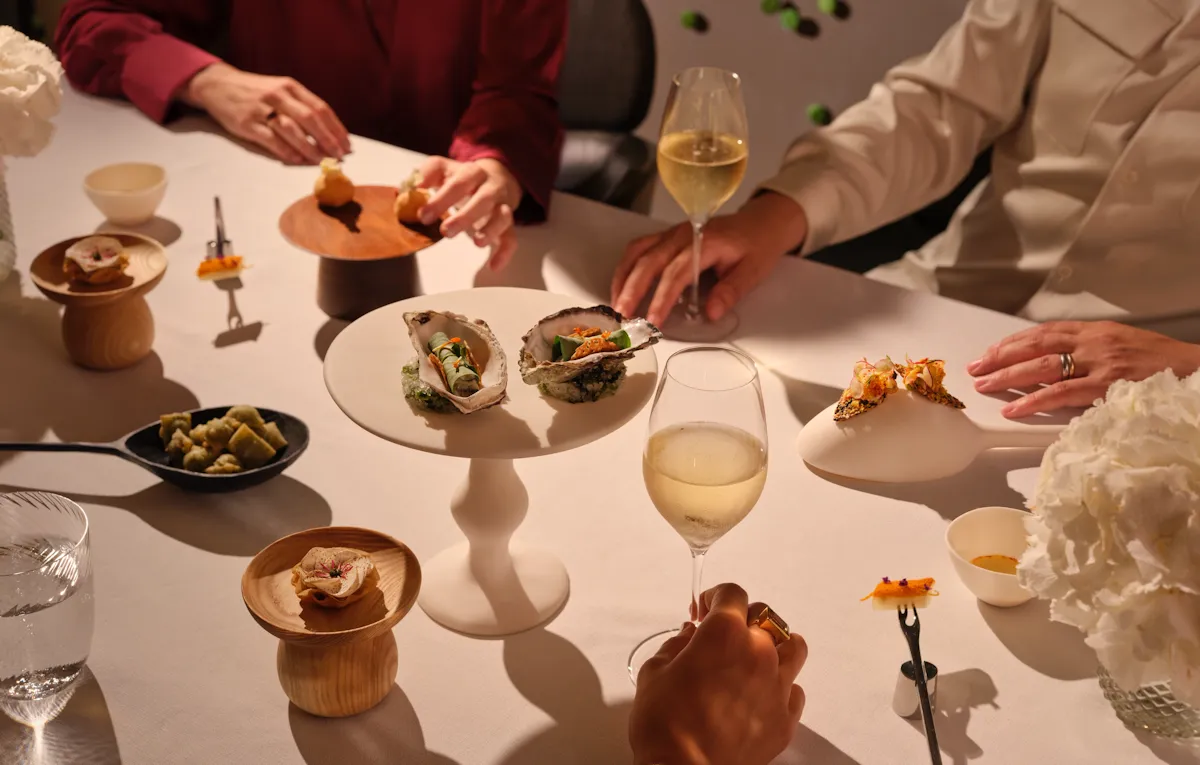 A beautifully styled dining table at Alain Ducasse at The Dorchester, set with a selection of refined tasting dishes served on sculptural tableware, accompanied by sparkling wine and white floral arrangements under warm ambient lighting.