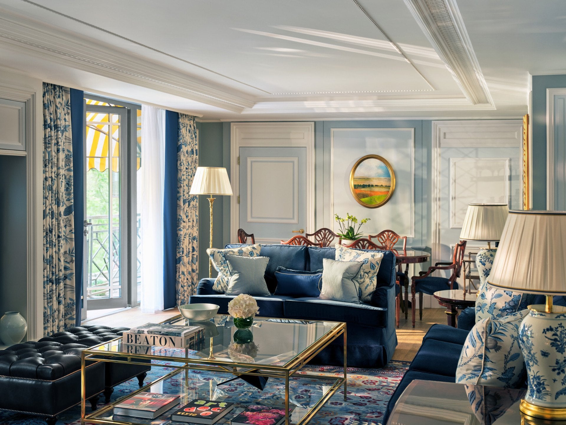 The Hyde Park Suite living area at The Dorchester, showcasing soft blue interiors, plush seating, decorative panelling, a glass coffee table and French doors opening onto a balcony with views of Hyde Park.
