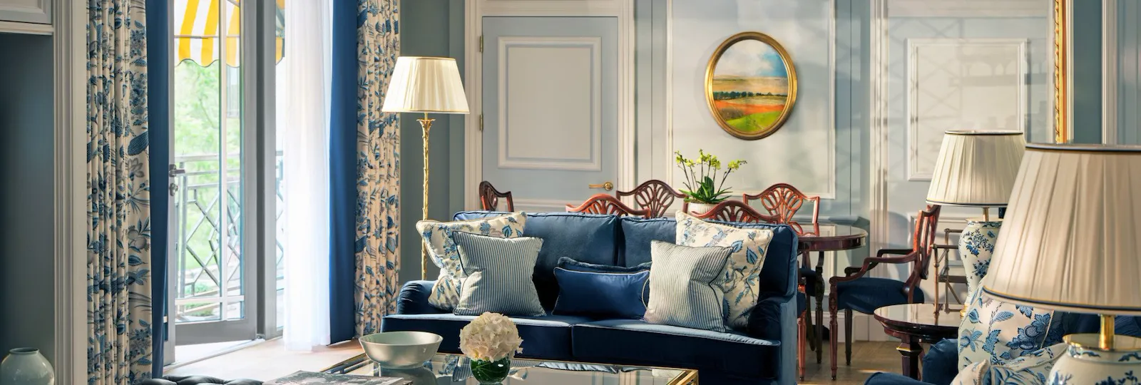 The Hyde Park Suite living area at The Dorchester, showcasing soft blue interiors, plush seating, decorative panelling, a glass coffee table and French doors opening onto a balcony with views of Hyde Park.