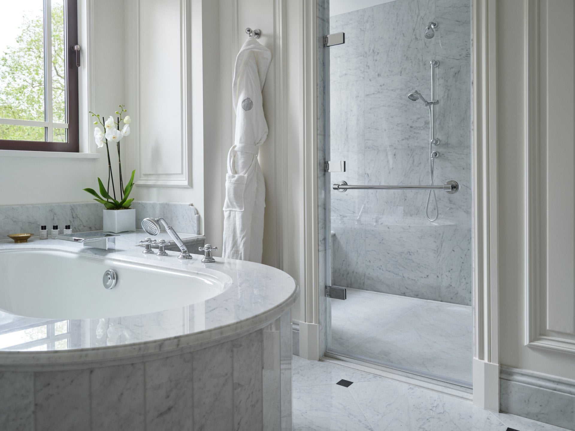 The Hyde Park Suite bathroom at The Dorchester, showcasing a curved marble bathtub, a spacious walk-in shower, white marble finishes and natural light from a large window.