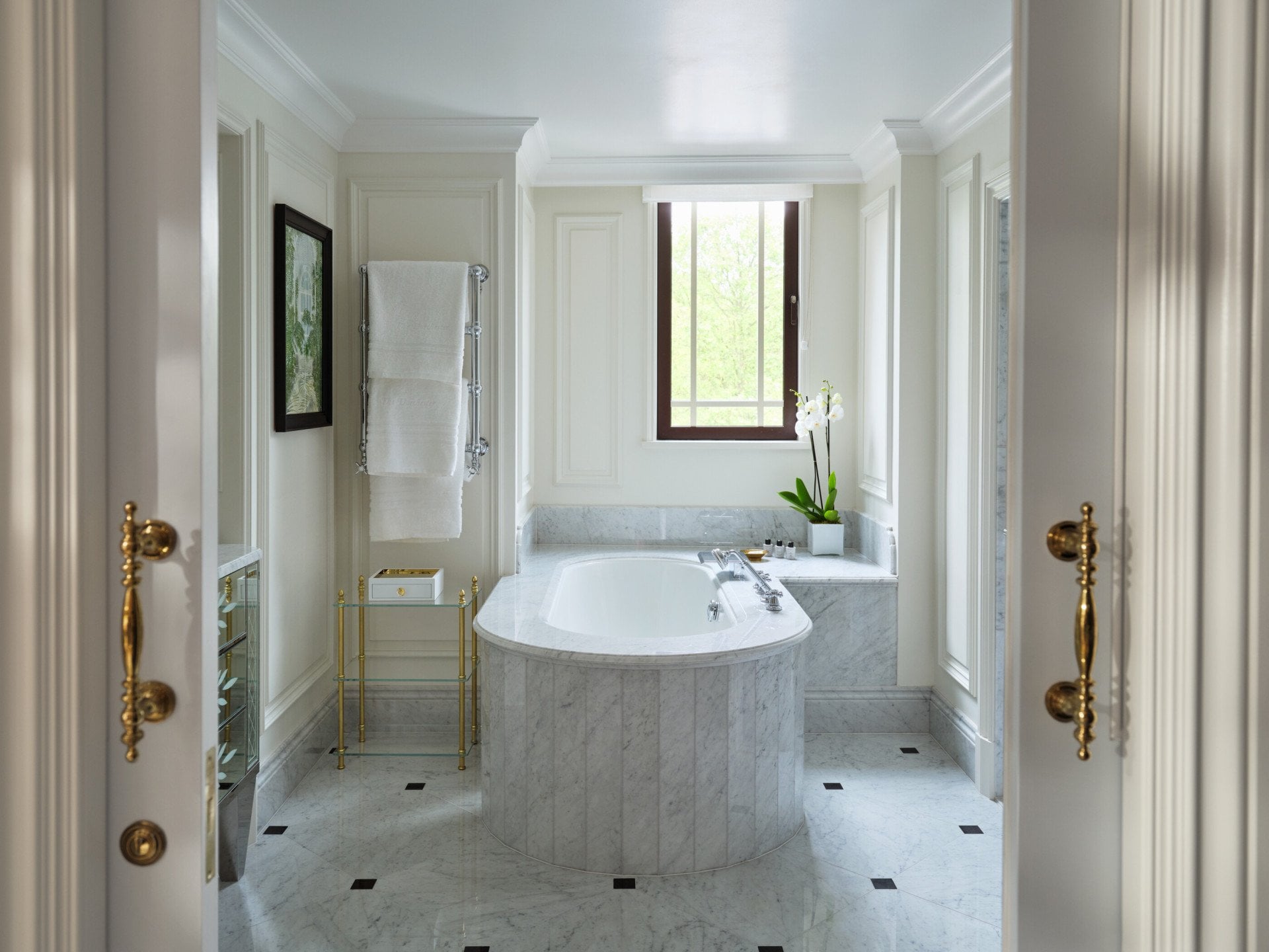 The Hyde Park Suite bathroom at The Dorchester, showcasing a freestanding oval bathtub, white marble finishes, gold detailing and a large window bringing in natural light.