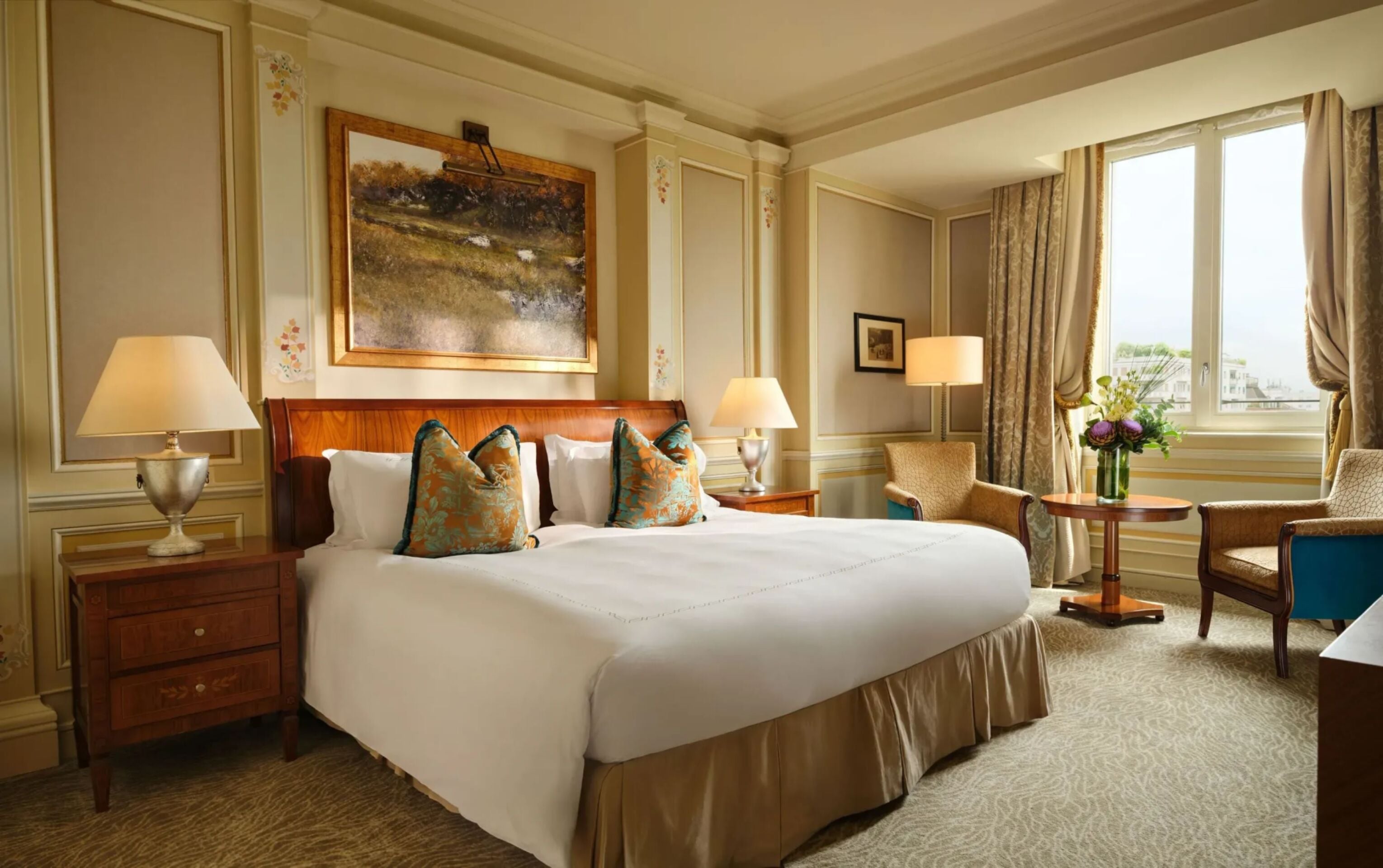 The room is decorated in soft gold colours and features decorative gold and blue cushions. There is a large painting above the bed and a large window that lets in plenty of natural light.