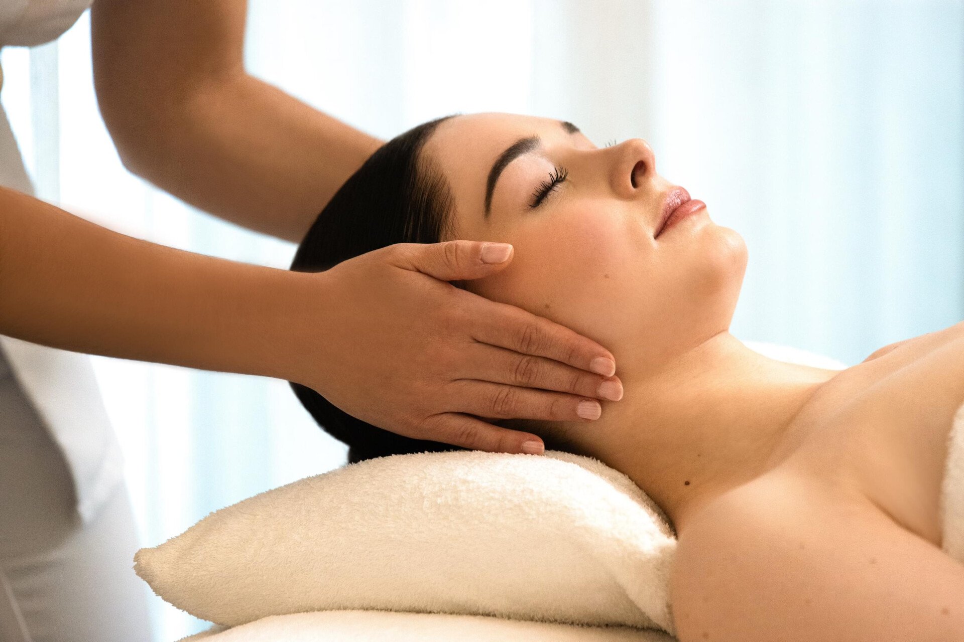 We can see a woman lying on a massage table, resting her head on a white pillow. We can see her from the side. The masseuse's hands are placed on the client's ears. The client's eyes are closed at the spa Valmont at Le Meurice, Paris.