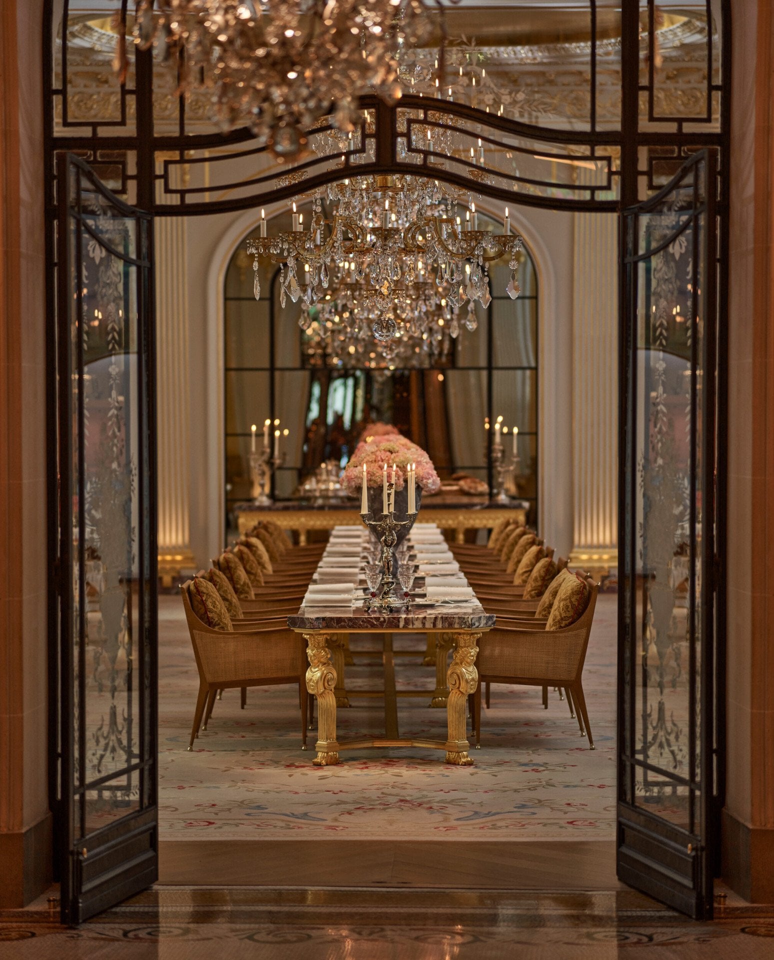 Jean Imbert au Plaza Athénée's central marble table, drawned up with pink flowers and chandeliers. Crystal chandeliers hanging above the table. Wrought iron and glass door at the forefront. Hotel Plaza Athénée, Paris