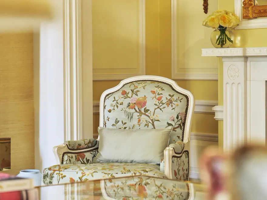 The Mayfair Suite at The Dorchester, showcasing a floral-patterned armchair, a glass-topped coffee table, soft yellow walls, a decorative fireplace and fresh cream roses displayed beneath a gilded mirror.