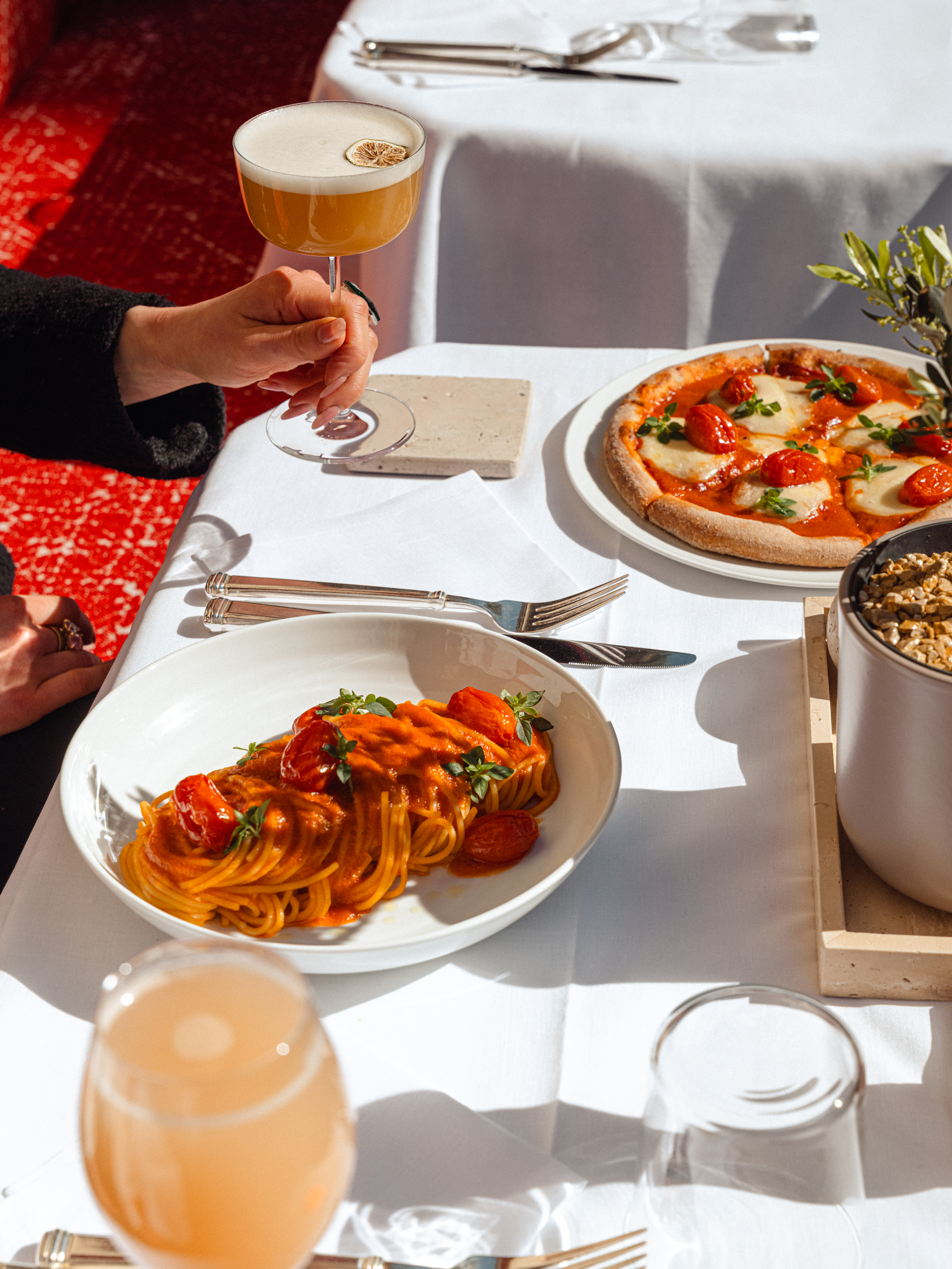 Pasta and pizza on a white table, a hand hold a cocktail. 