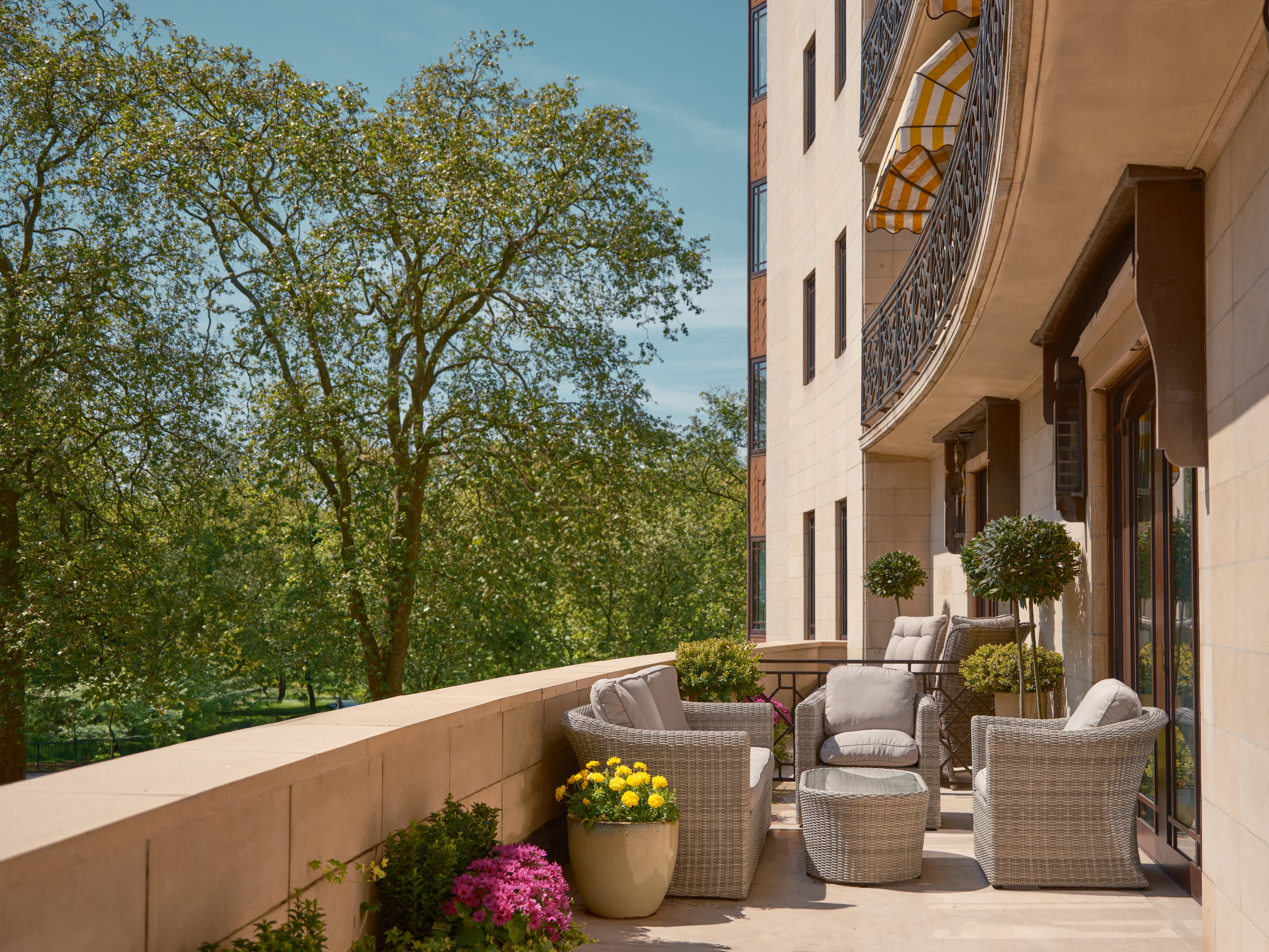 Spacious private terrace at The Dorchester’s Garden Terrace Suite, with woven armchairs and tables arranged for outdoor relaxation, framed by colourful potted flowers and overlooking a tranquil canopy of trees on a bright summer day.