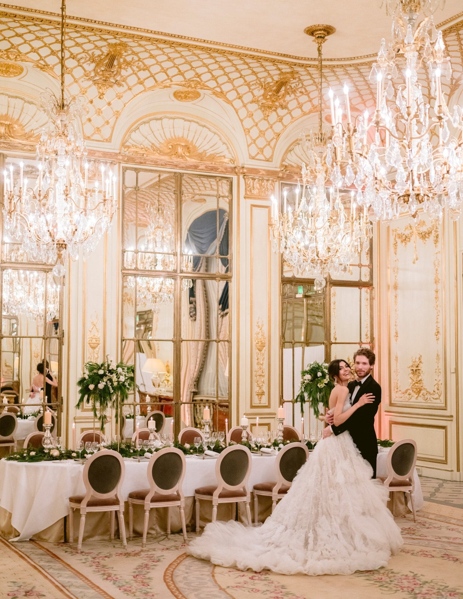 Wedding couple standing in front of a festive set table in Salon Pompadour. Green and white flower arrangements are on the table. The bride is laughing and in the arms of her husband, at Le Meurice, Paris