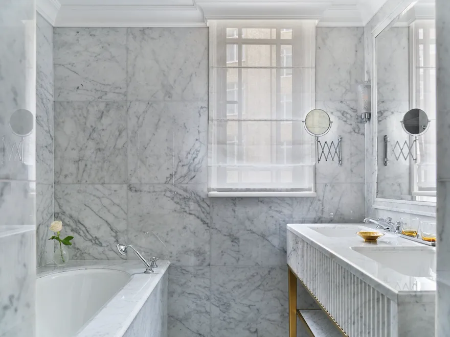 The Superior Room bathroom at The Dorchester, showcasing floor-to-ceiling white marble, a built-in bathtub, a fluted marble double vanity and a large mirror that enhances the bright, airy space.
