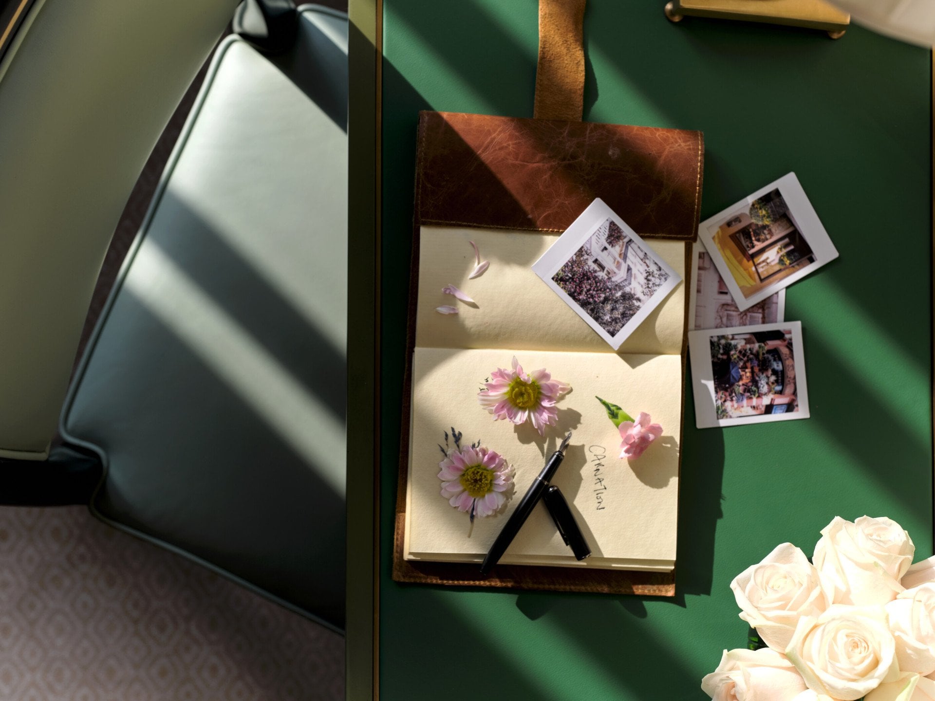 Detail of the Executive Room at The Dorchester, showcasing a green writing desk with pressed botanical artwork, instant photographs, a pen and a bouquet of white roses illuminated by natural light.