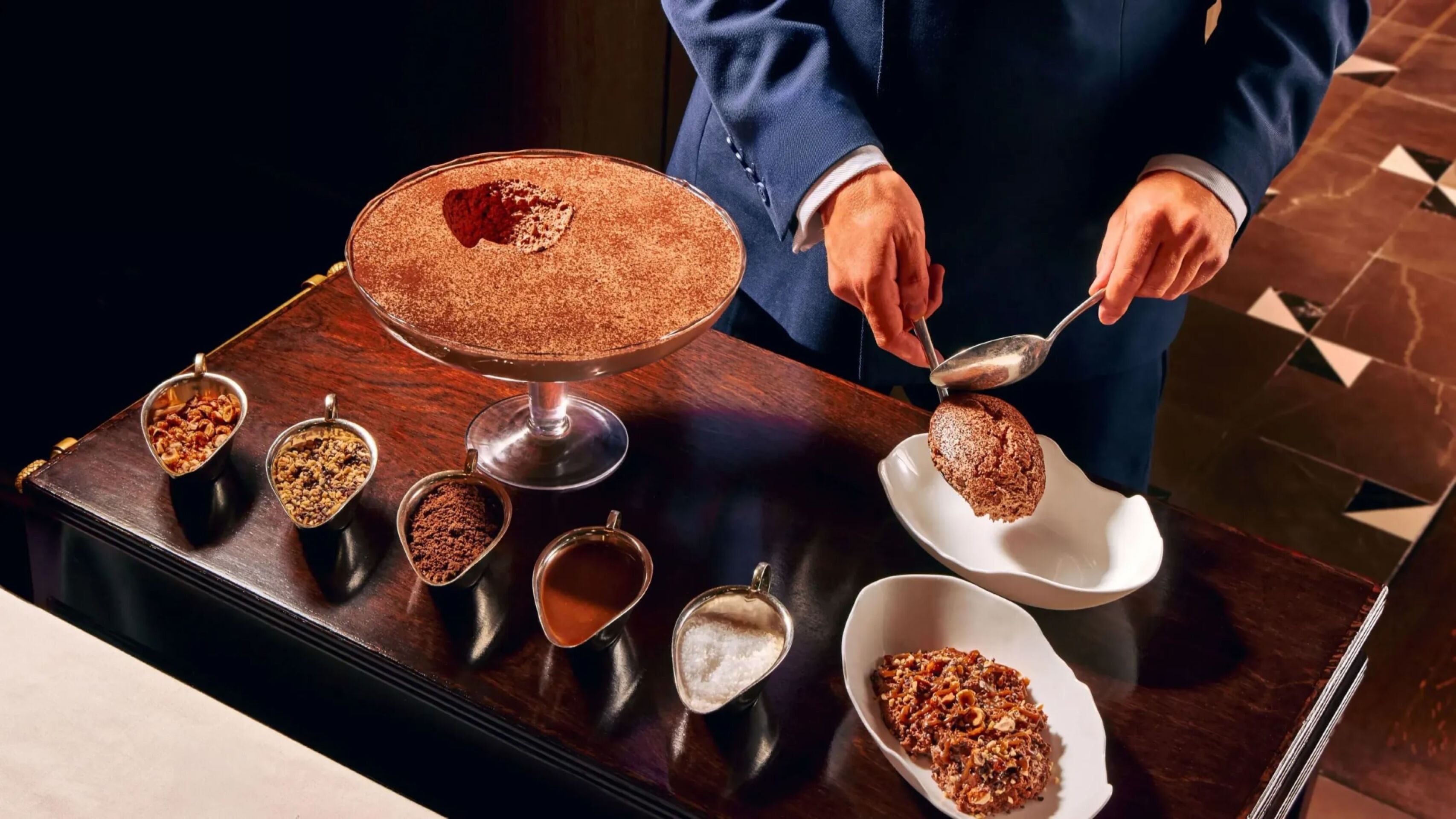 Tableside service of chocolate mousse at The Grill at The Dorchester, featuring a server plating rich chocolate mousse from a large sharing dish alongside an assortment of toppings including nuts and chocolate.