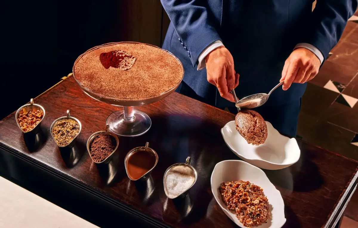 Tableside service of chocolate mousse at The Grill at The Dorchester, featuring a server plating rich chocolate mousse from a large sharing dish alongside an assortment of toppings including nuts and chocolate.