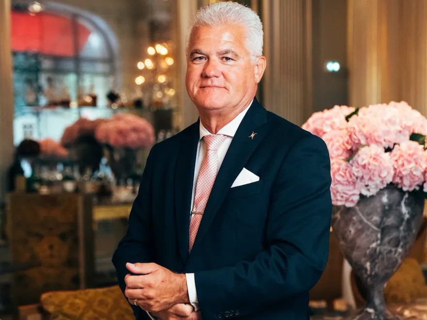 Person in formal suit standing in elegant room with pink flower arrangement and marble table.
