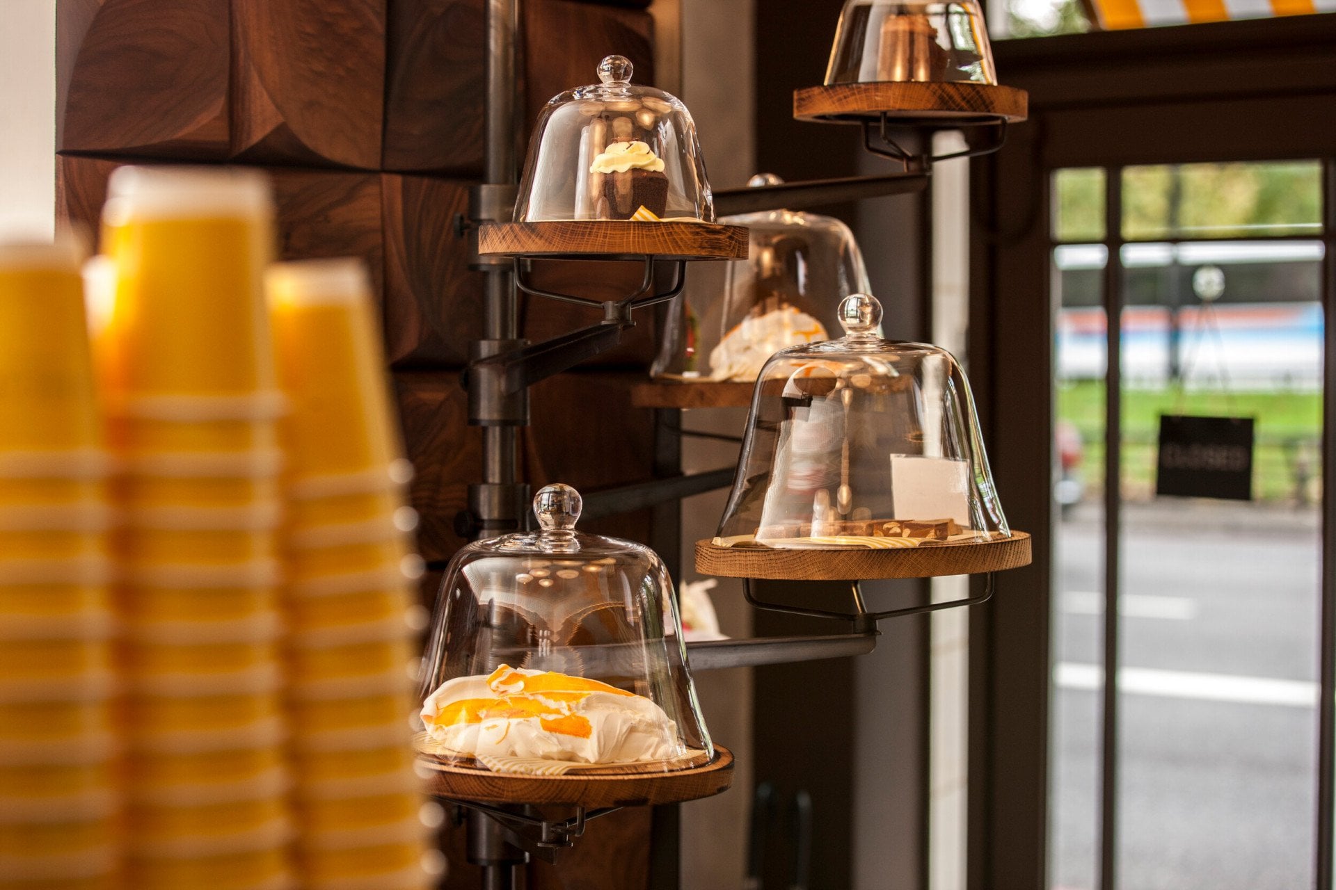 Display of freshly prepared pastries at Parcafé at The Dorchester, showcasing sweet treats presented under glass cloches on tiered shelves, with takeaway coffee cups and views towards Hyde Park visible through the café window.