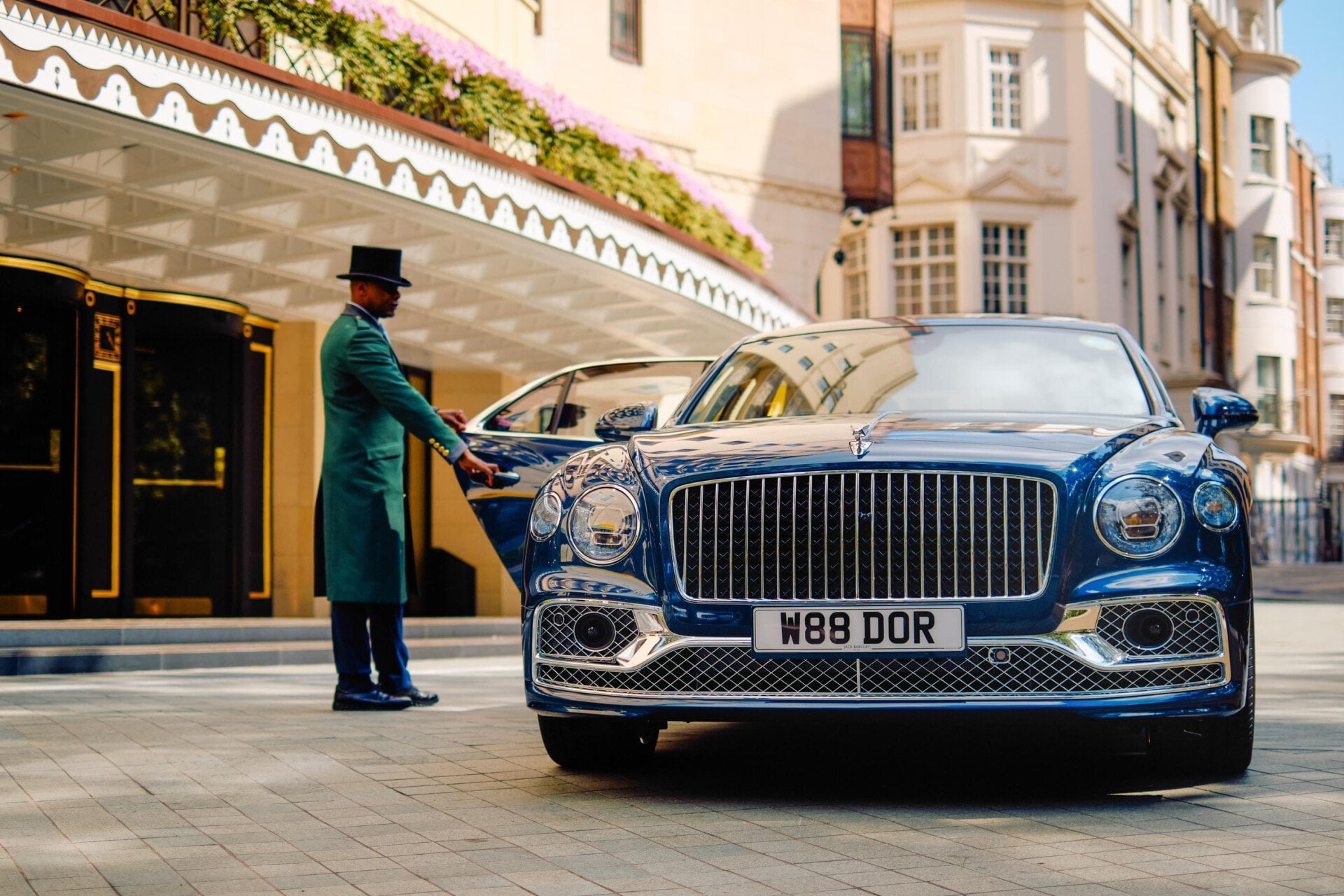 The Dorchester Bentley positioned outside the hotel's main entrance on Park Lane, showcasing its distinctive chrome grille and personalised registration plate, with The Dorchester's elegant façade and entrance canopy in the background.