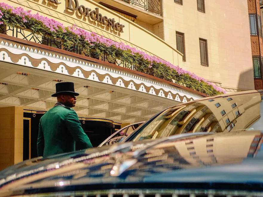 The Dorchester Bentley parked outside the hotel's main entrance on Park Lane, showcasing its distinctive grille and polished finish against the backdrop of The Dorchester's elegant façade, floral displays and signature signage.