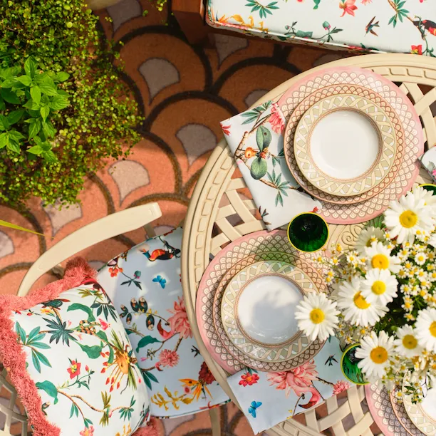 The Dorchester La DoubleJ Summer Garden At The Dorchester. Outdoor table setting with floral cushions, patterned tableware and fresh daisies on a tiled terrace surrounded by greenery.