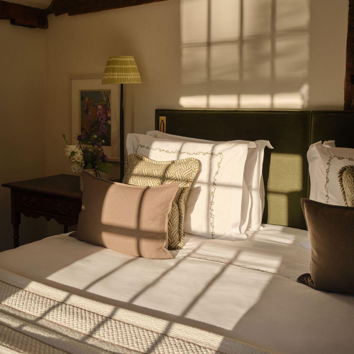 Sunlit bedroom with exposed beams and green velvet headboard in a cottage room at Coworth Park, Ascot