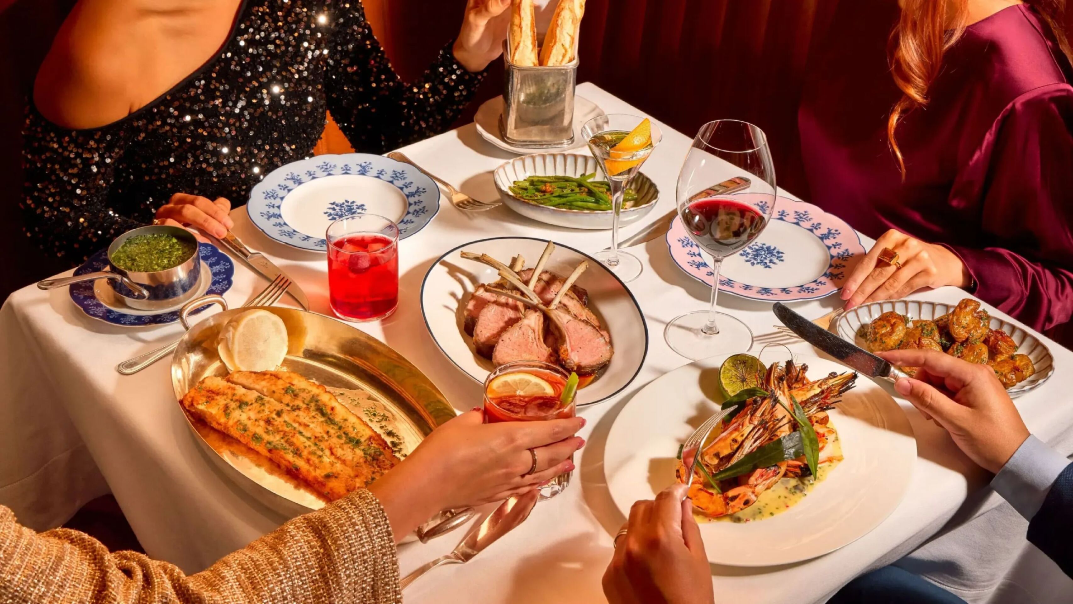 Overhead view of a dining table at The Grill at The Dorchester with lamb chops, grilled prawns, Dover sole, green beans, sauces, cocktails and guests serving themselves from shared dishes.
