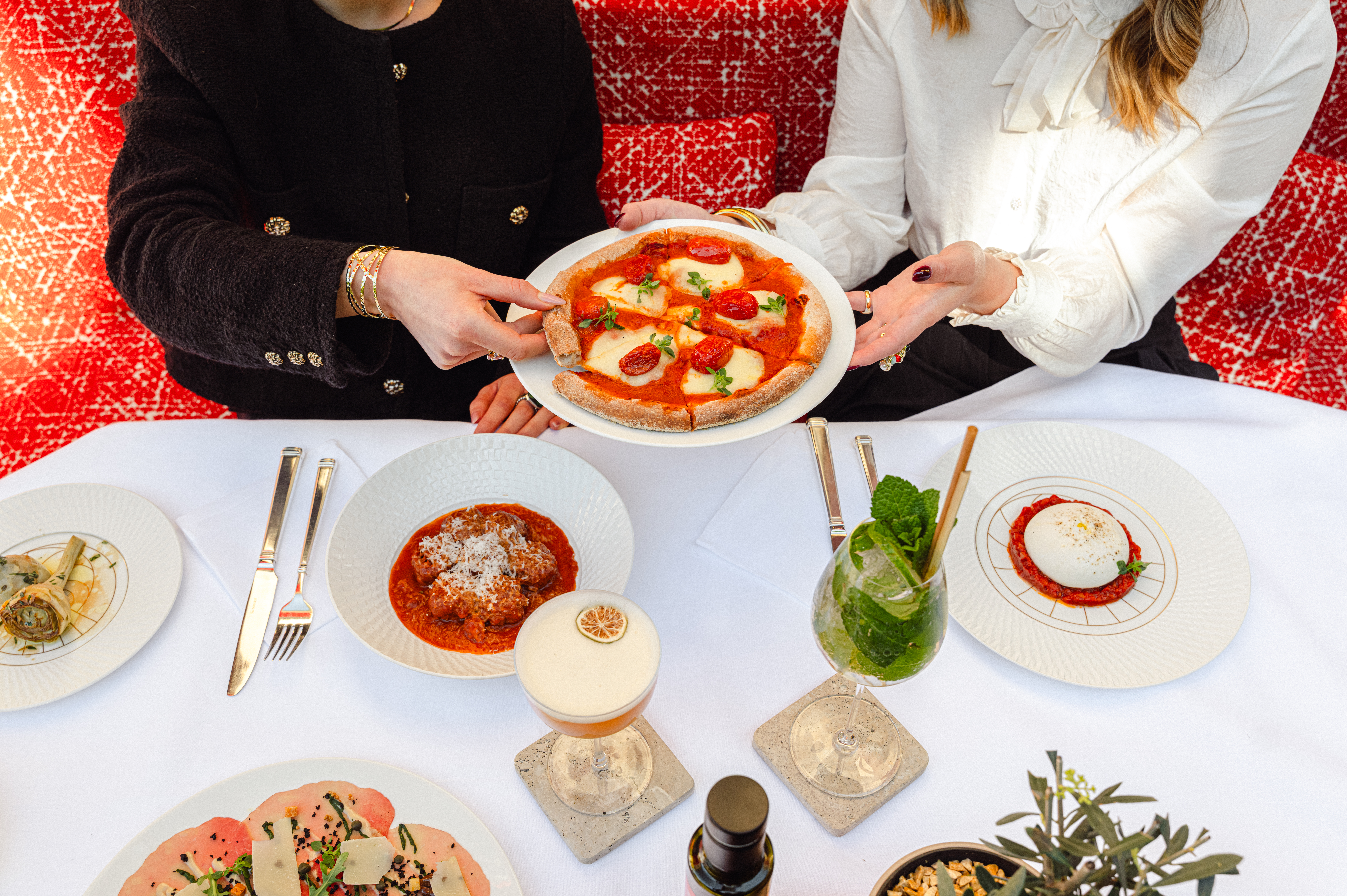 Two woman sitting on a red bench sharing a pizza, cocktails and Italian dishes are displayed on a white table. 