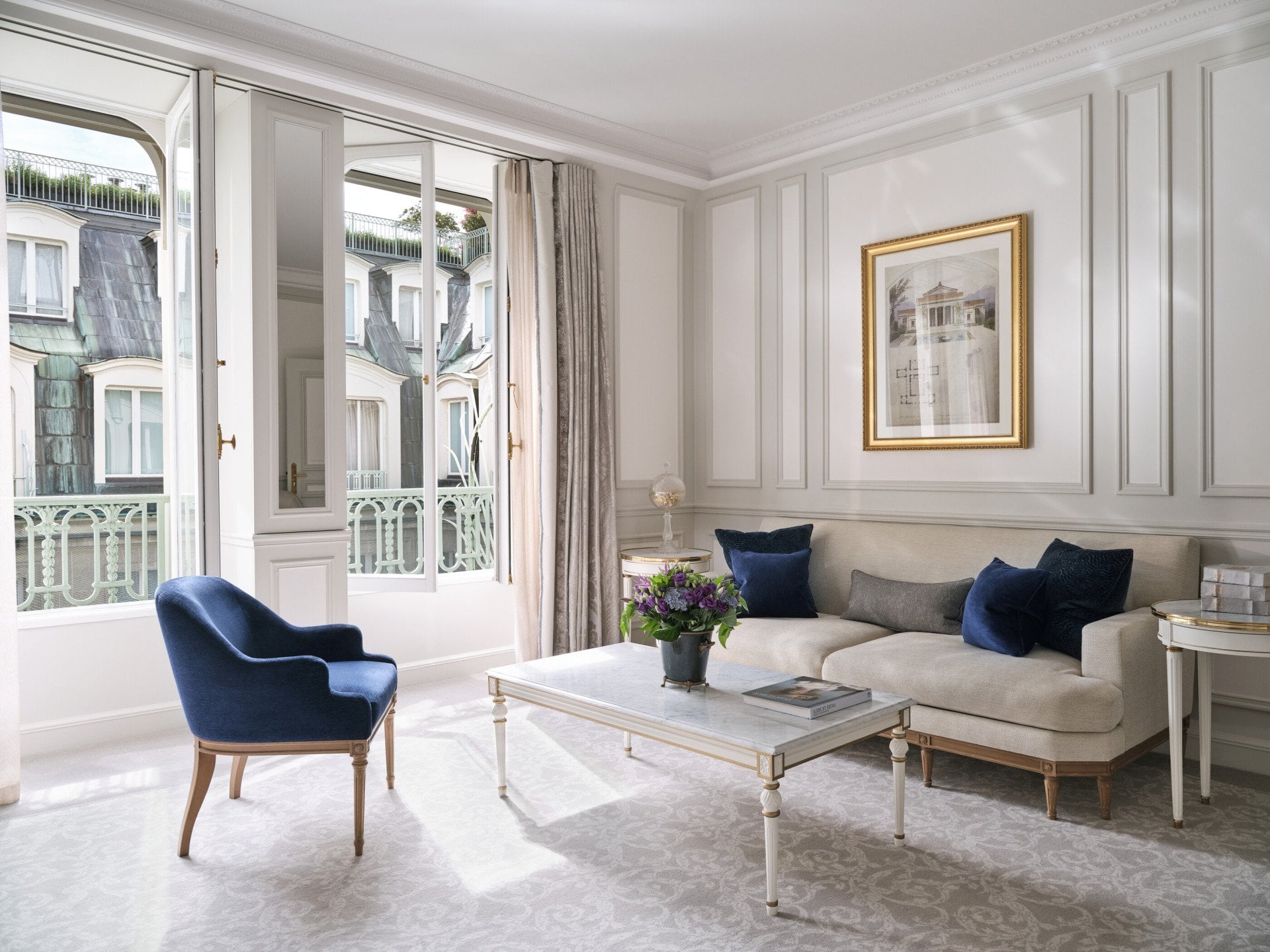 Living room of a Deluxe Suite with a chair, a painting above the couch and a table with flowers and books on the top, at Le Meurice, Paris
